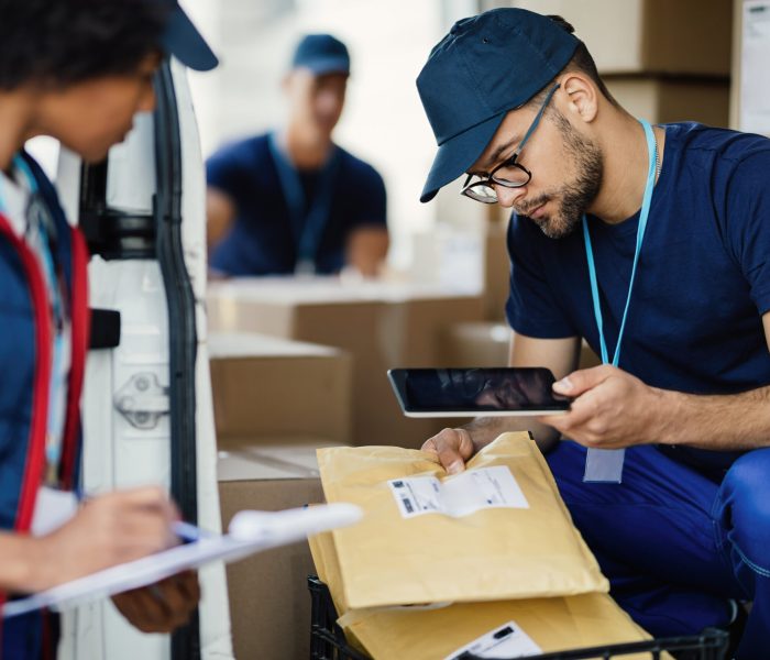 Young delivery man using digital tablet for scanning bar code on package label while preparing for shipment with his coworker.